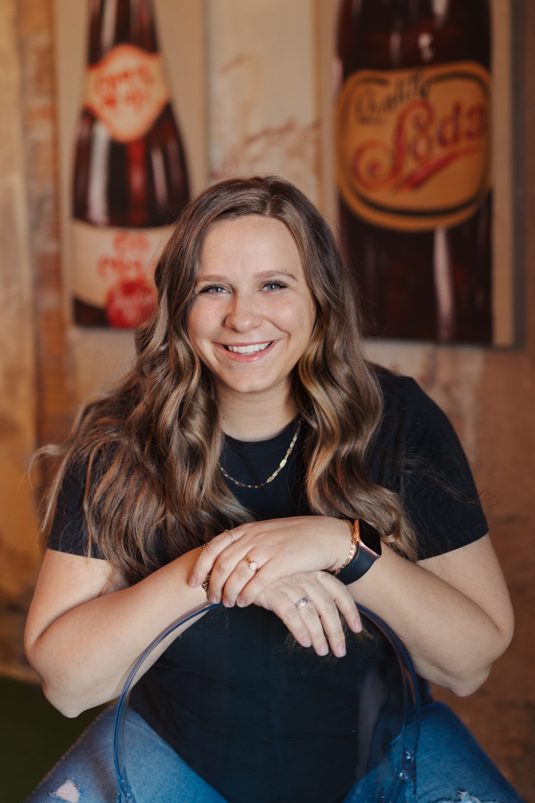 Marisa Grover smiling, leaning on the back of a chair in front of vintage soda signs