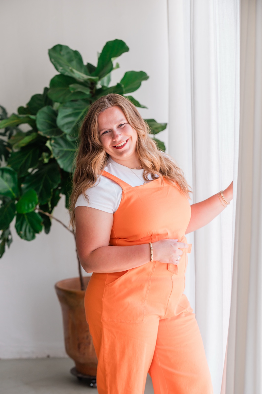 Marisa Grover smiling in orange overalls next to a fiddle leaf fig